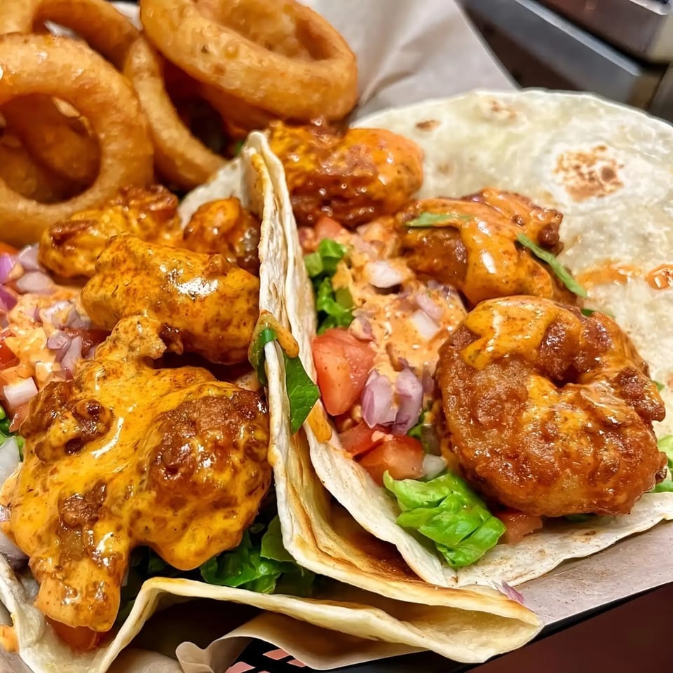 Two shrimp tacos with lettuce, tomato, onion, and sauce, served with a side of onion rings.
