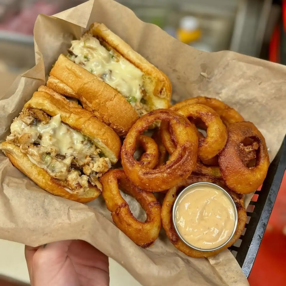 Two cheesy chicken cheesesteak sandwiches and a pile of crispy onion rings with dipping sauce.